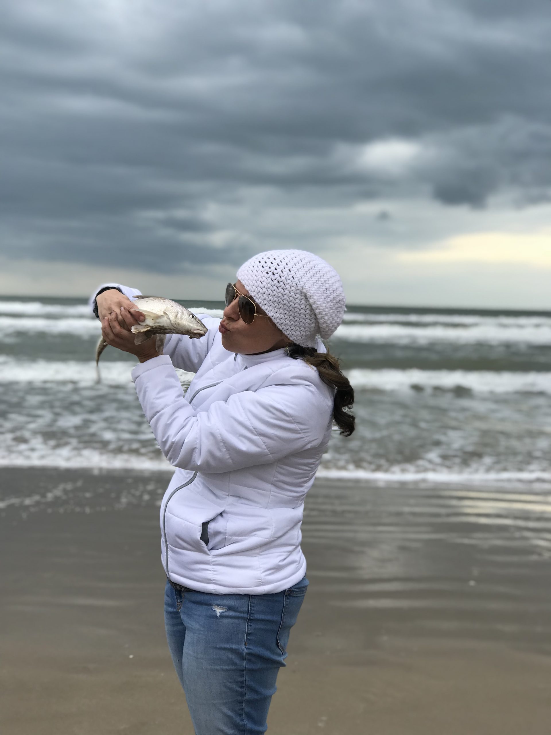 Photo of an ominous, dark and cloudy sky. On the beach. Waves crash upon the shore. Standing on the shore, a woman in jeans, a white puffer coat, white knit hat, and aviator sunglasses. She is holding a fish up to close to her face, about to kiss it!