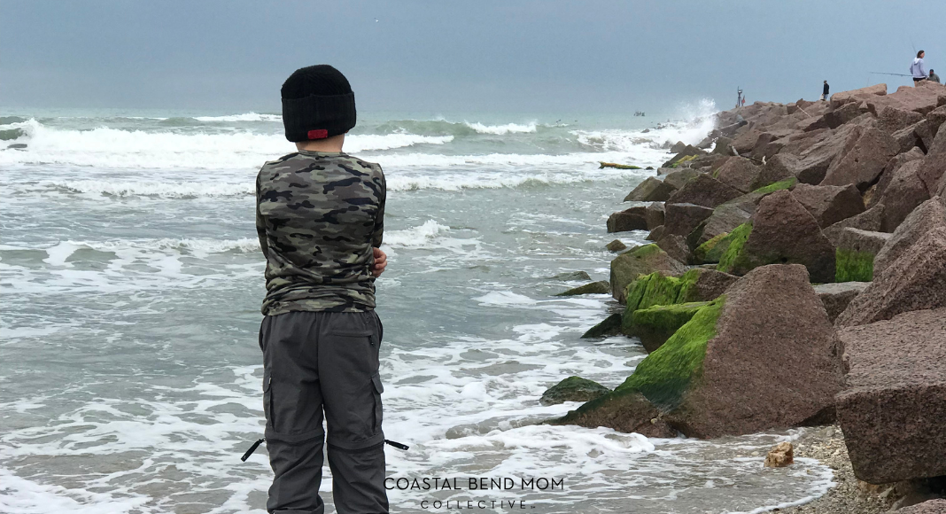 Boy in a black winter hat, long sleeves, and pants, hugging himself, looking out at the cold ocean. Large granite boulders create a jetty on the right hand side. Waves are crashing against the jetty.