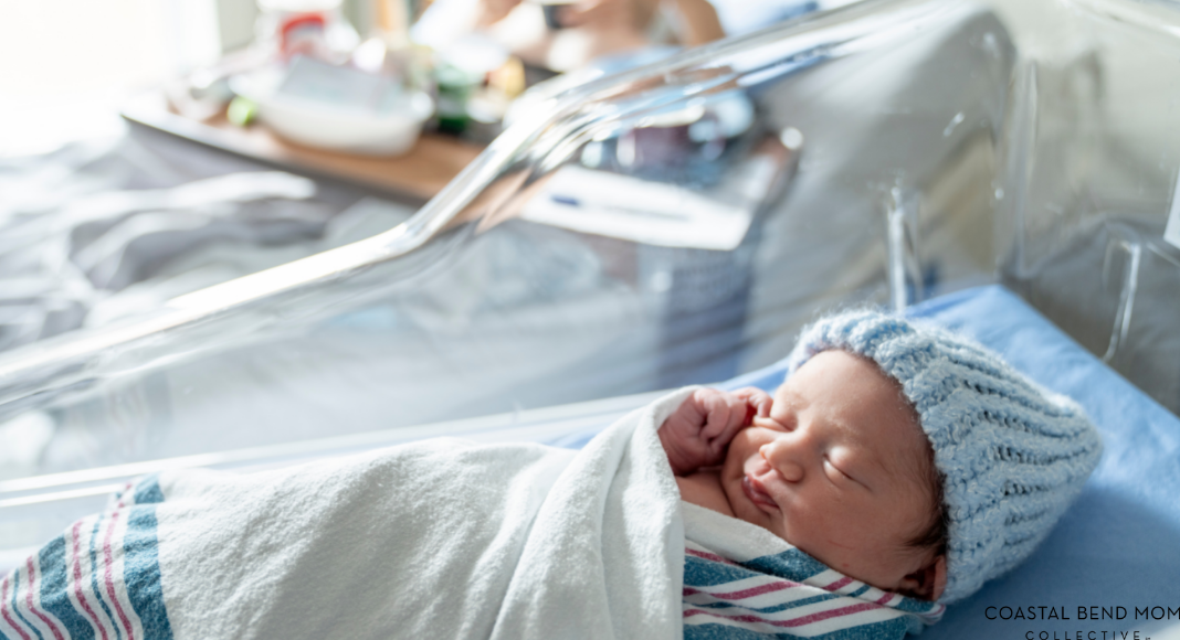 Image of a newborn baby, laying in a bassinet, wrapped in a receiving blanket wearing a knitted blue hat. In the background you can see the new mom laying in the hospital bed.