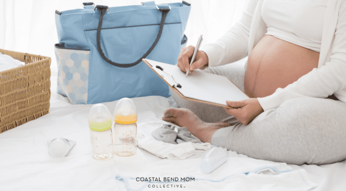 Image of an expecting mom, sitting cross legged on a white bed, making a list on a clipboard, planning things to take to the hospital. Also pictured: a blue diaper bag, 2 baby bottles, and a pacifier.