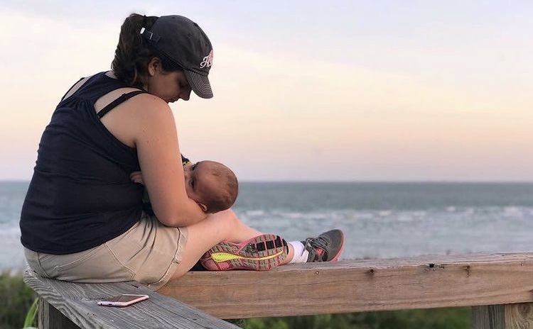 Image of a mom wearing a black tank top, khaki shorts, and a black baseball cap with a ponytail. She is sitting on a wooden bench, at sunset, with a view of the ocean in the background. She is nursing an infant boy.