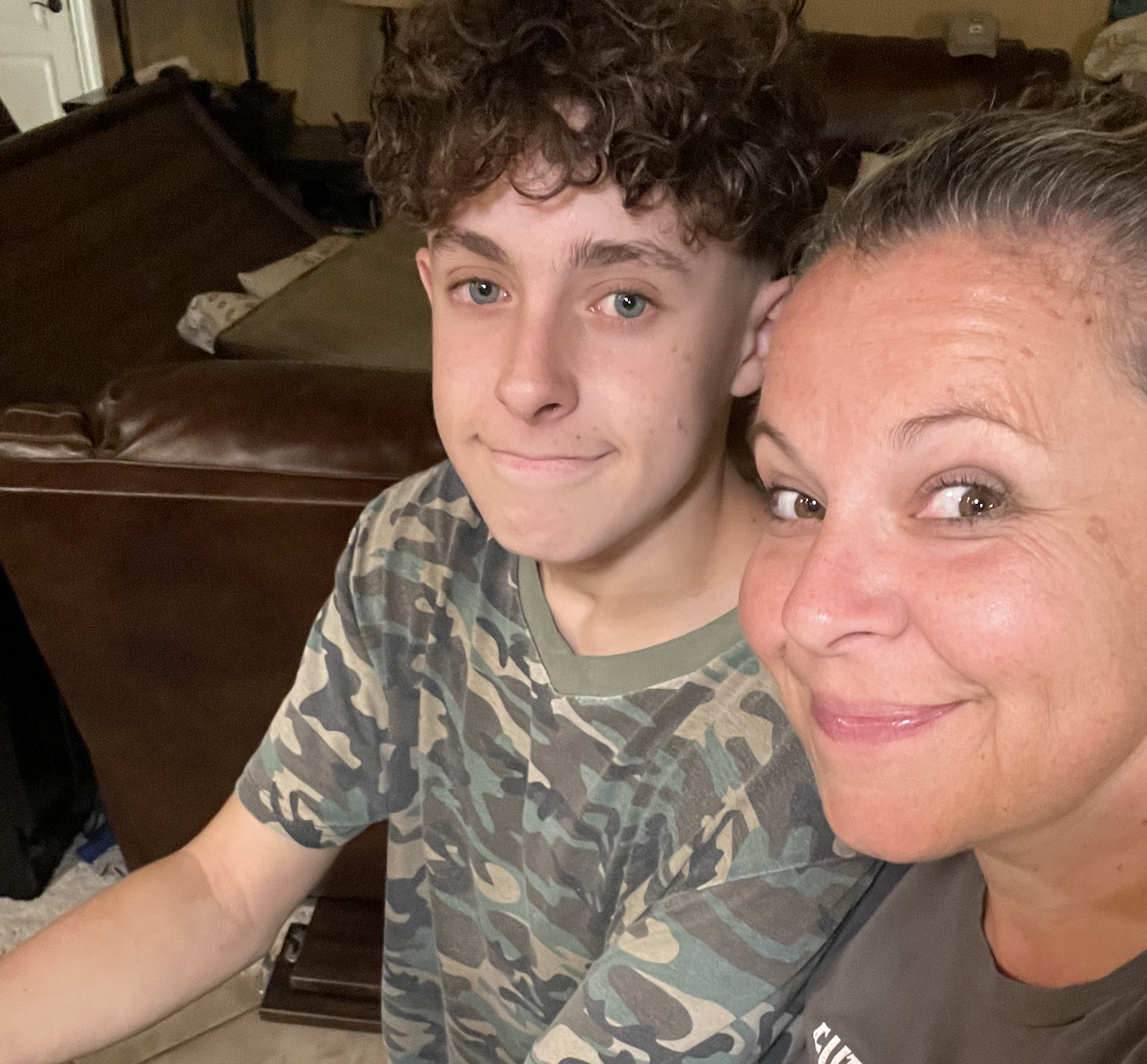 Image of a teenage boy in a camouflage t-shirt, smiling without showing teeth. Also pictured, sitting next to him, his mom, also smiling with no teeth.