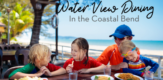 Image: A mom wearing an orange shirt and blue baseball cap, is eating lunch with three children. From left to right, there is a blond haired boy wearing a green shirt, he is talked to a brunette little girl, wearing a red shirt. Sitting on mom's lap is a child wearing goggles and a rainbow striped sun shirt. They are eating lunch with the ocean behind them.