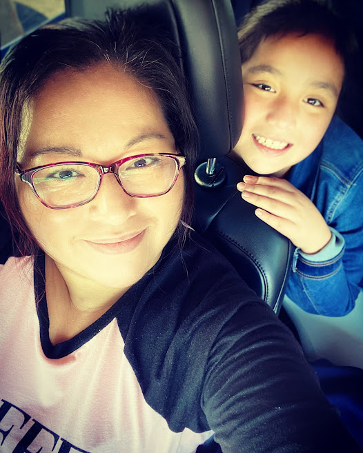 Image of a Latina mother and her daughter. The mother is sitting in the passenger seat of a car and wearing glasses. The daughter is sitting in the backseat, leaning forward, smiling a big smile.