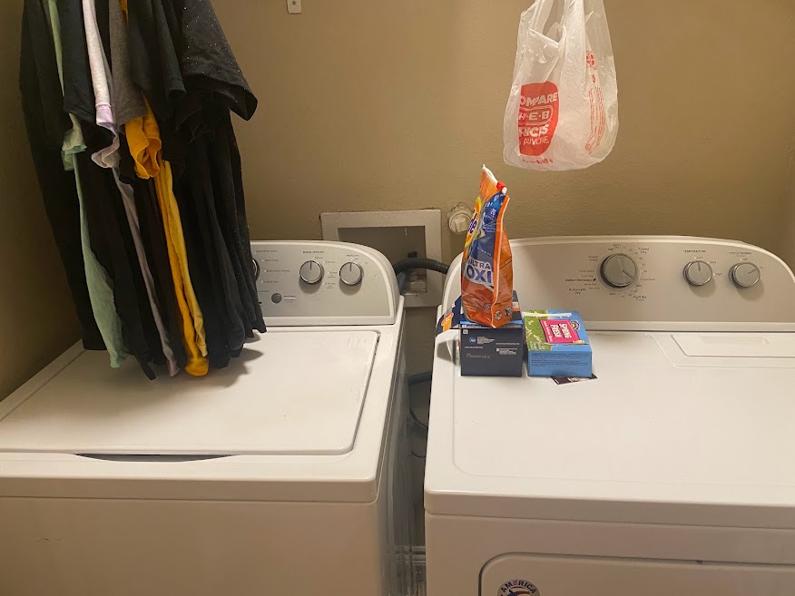 Image of a washing machine and a dryer. Hanging above the washing machine are several t-shirts hanging from hangers. Sitting on top of the dryer are two boxes of dryer sheets and a bag of washing machine detergent pods.