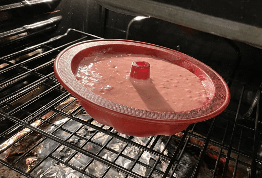 Image of a red bundt cake pan, filled with strawberry cake batter. Pan is sitting on the upper rack of the oven.