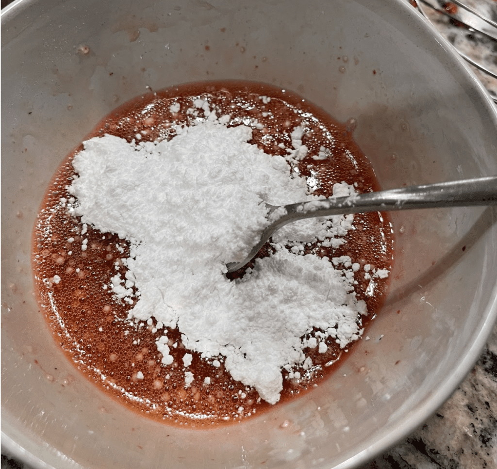 Image of a bowl of strawberry juice, with white powdered sugar being stirred in with a silver spoon.