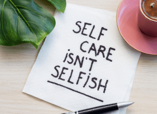 Image shows a green monstera leaf in the upper left corner, a pink teacup on a pink saucer in the upper right corner. In the center is a white napkin that has been written on in a black marker. It reads: Self Care Isn't Selfish