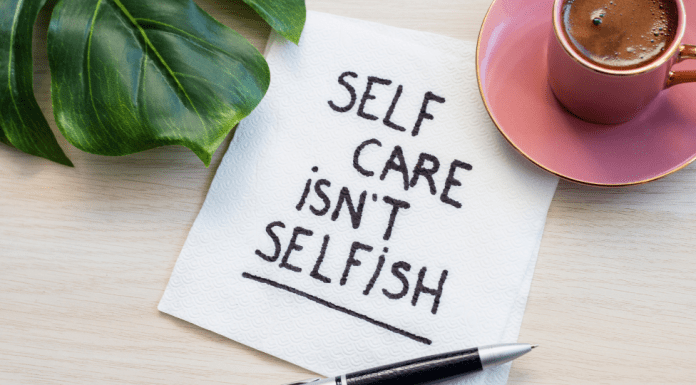 Image shows a green monstera leaf in the upper left corner, a pink teacup on a pink saucer in the upper right corner. In the center is a white napkin that has been written on in a black marker. It reads: Self Care Isn't Selfish
