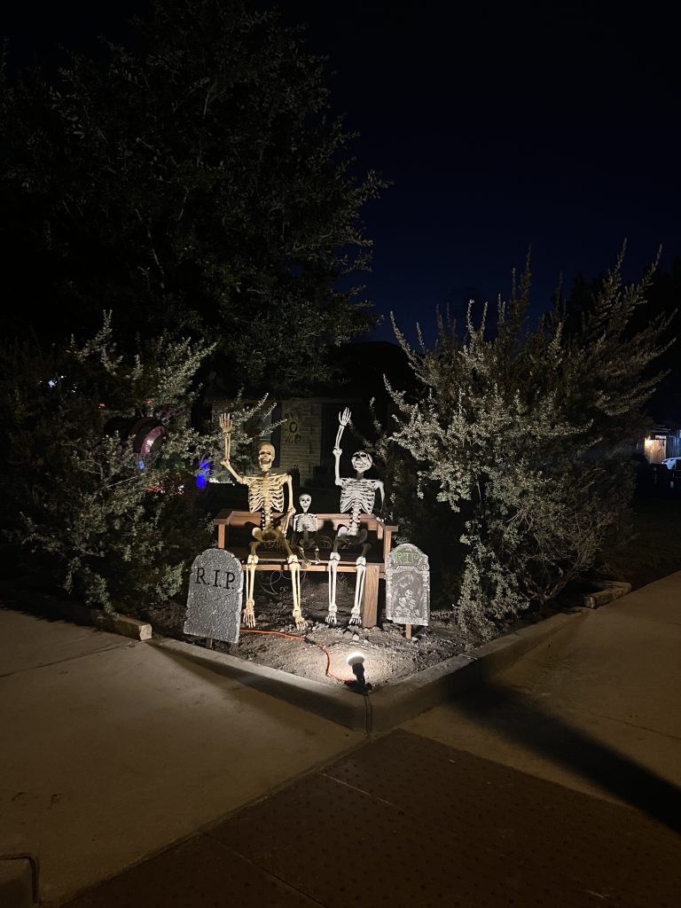 Halloween Decorations, outside of a house at night, uprights shibnging on fake tombstones and two skeletons