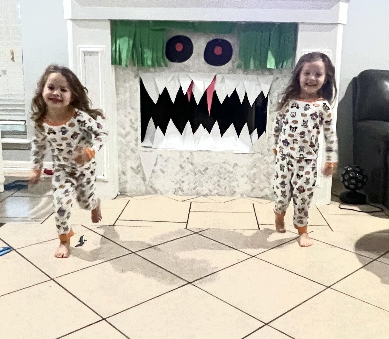 Boy Girl Twins in matching halloween pajamas, standing in front of a fireplace that has been transformed to look like a monster using paper to make green fringe hair, google eyes, and big white triangular fangs and teeth.