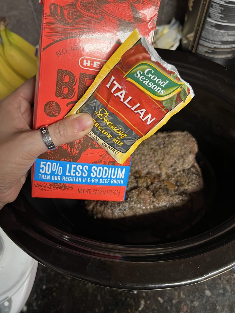 Image shows a seared pot roast inside of a crockpot. You see a woman's hand holding a box of beef bone broth and a packet of Italian dressing seasoning.