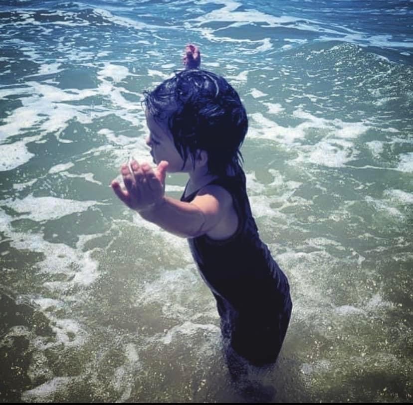 Image of a little boy with brown hair playing in the surf at the beach. He is wearing black swim trunks and a black tank top. He is holding his hands in the air. This is Jacob's Autism Story.