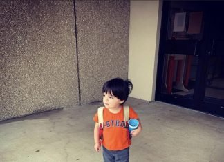 Image of a little boy with dark brown hair. He is taking a step forward, wearing blue shorts and an orange Astros t-shirt. He's holding a blue sippy cup. This is Jacob's Autism Story.