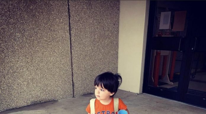 Image of a little boy with dark brown hair. He is taking a step forward, wearing blue shorts and an orange Astros t-shirt. He's holding a blue sippy cup. This is Jacob's Autism Story.