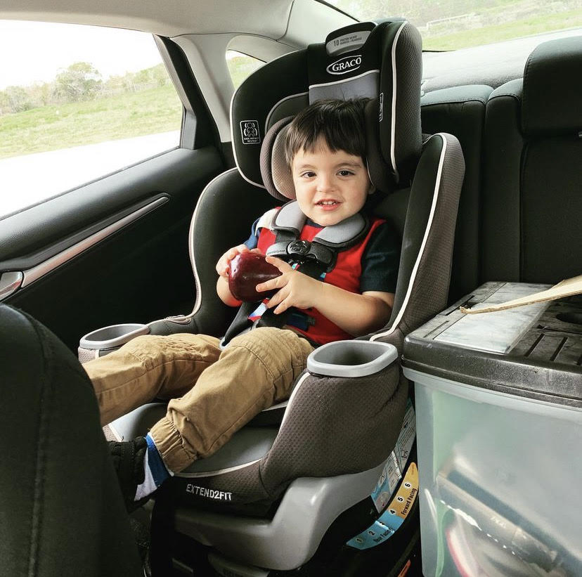 Image of a toddler boy with dark brown hair. He is sitting in a car seat, buckled in, smiling towards the camera. He has on khaki pants and a red t-shirt with navy blue sleeves.