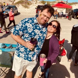Man in a blue Hawaiian shirt and khaki shorts, holding a cup. Has his arm around a woman in sunglasses holding a pink tumbler. They are at the beach attending Barefoot Mardi Gras.