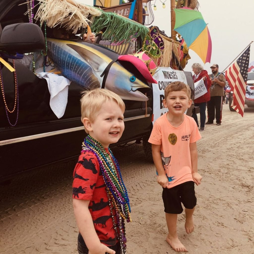 Image of two little boys, standing in front of a Mardi Gras parade float. One of the boys has Mardi grad beads around his neck. They are barefoot in the sand.