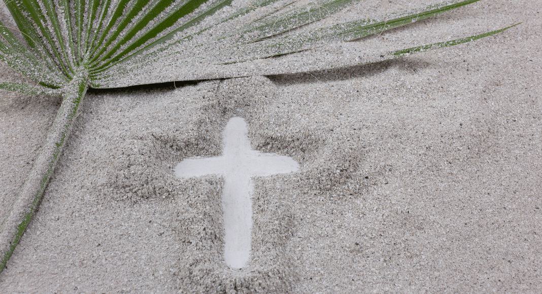 Image of a cross in the sand. In the upper left corner a palm branch lays in the sand.
