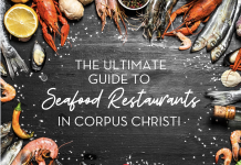 Black slate background, with an assortment of fresh seafood items creating a border. The text in the center reads "The Ultimate Guide to Seafood Restaurants in Corpus Christi"